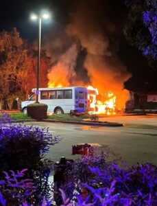1 bus fully engulfed with flames next to another parked bus in a parking lot.